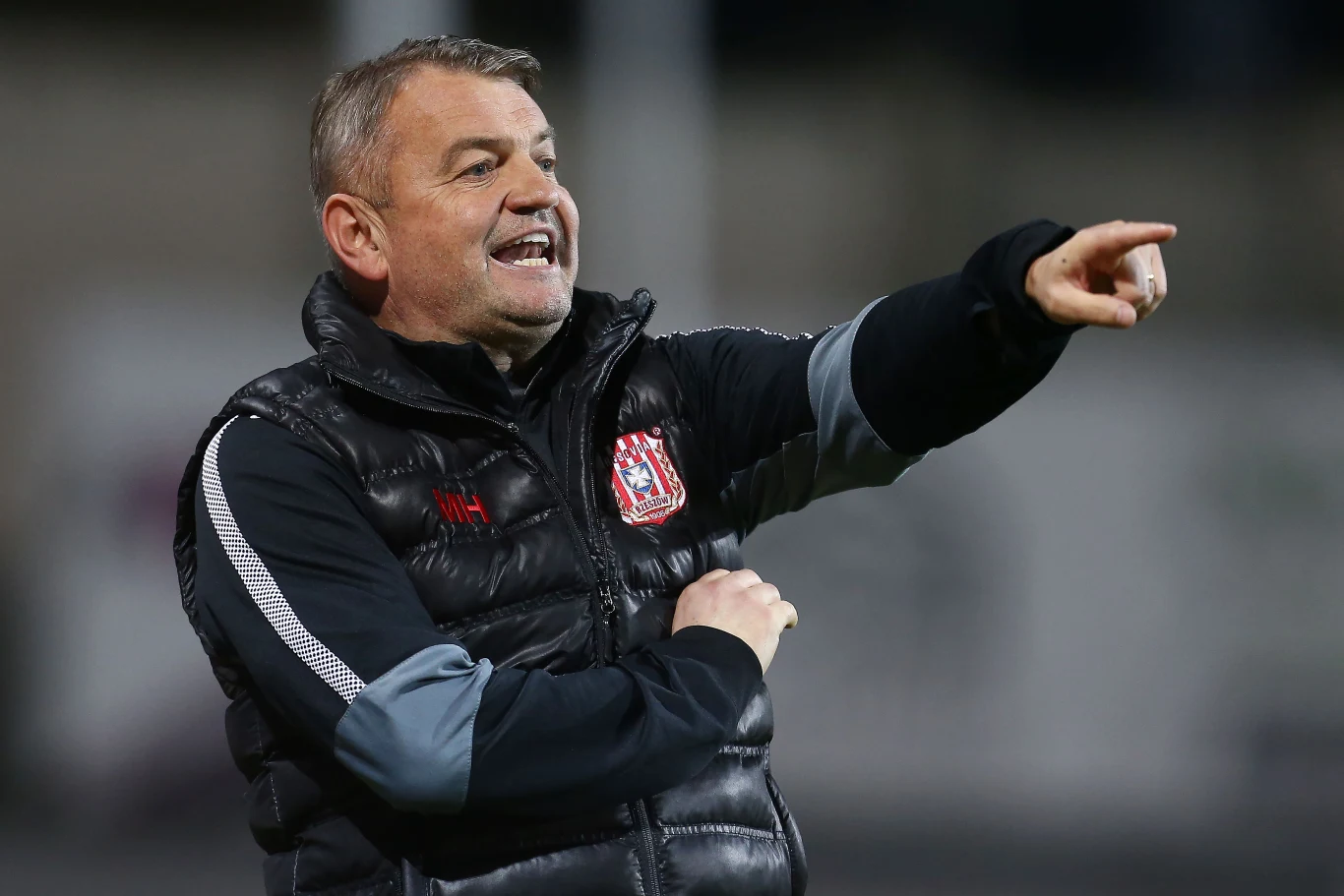 Mężczyzna w czarnej, pikowanej kurtce z herbem klubu sportowego energicznie wskazuje palcem w bok, prawdopodobnie wydając polecenia na boisku.