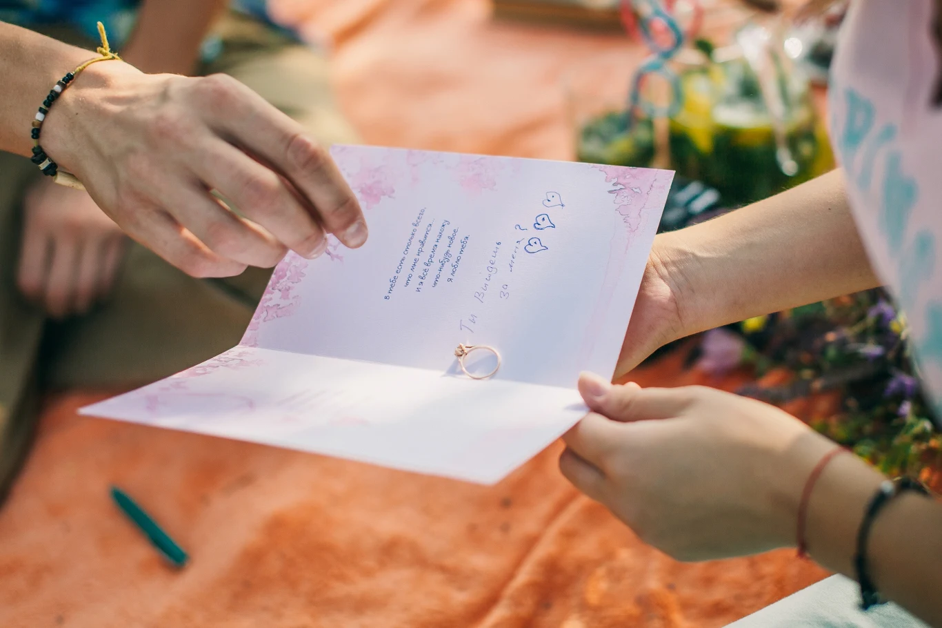 Dwie osoby trzymają otwartą kartkę z życzeniami, na której znajduje się pierścionek, wokół widać kwiaty oraz kolorowe dekoracje.