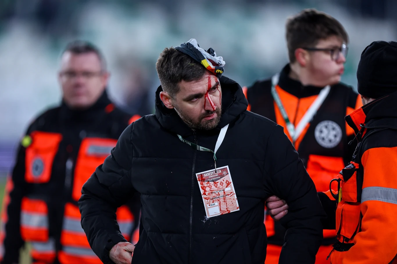 Mężczyzna z krwawiącą raną na głowie, opatrzony przez ratowników medycznych w jaskrawopomarańczowych kurtkach, na jego szyi zawieszona identyfikacja, atmosfera napięcia i troski.