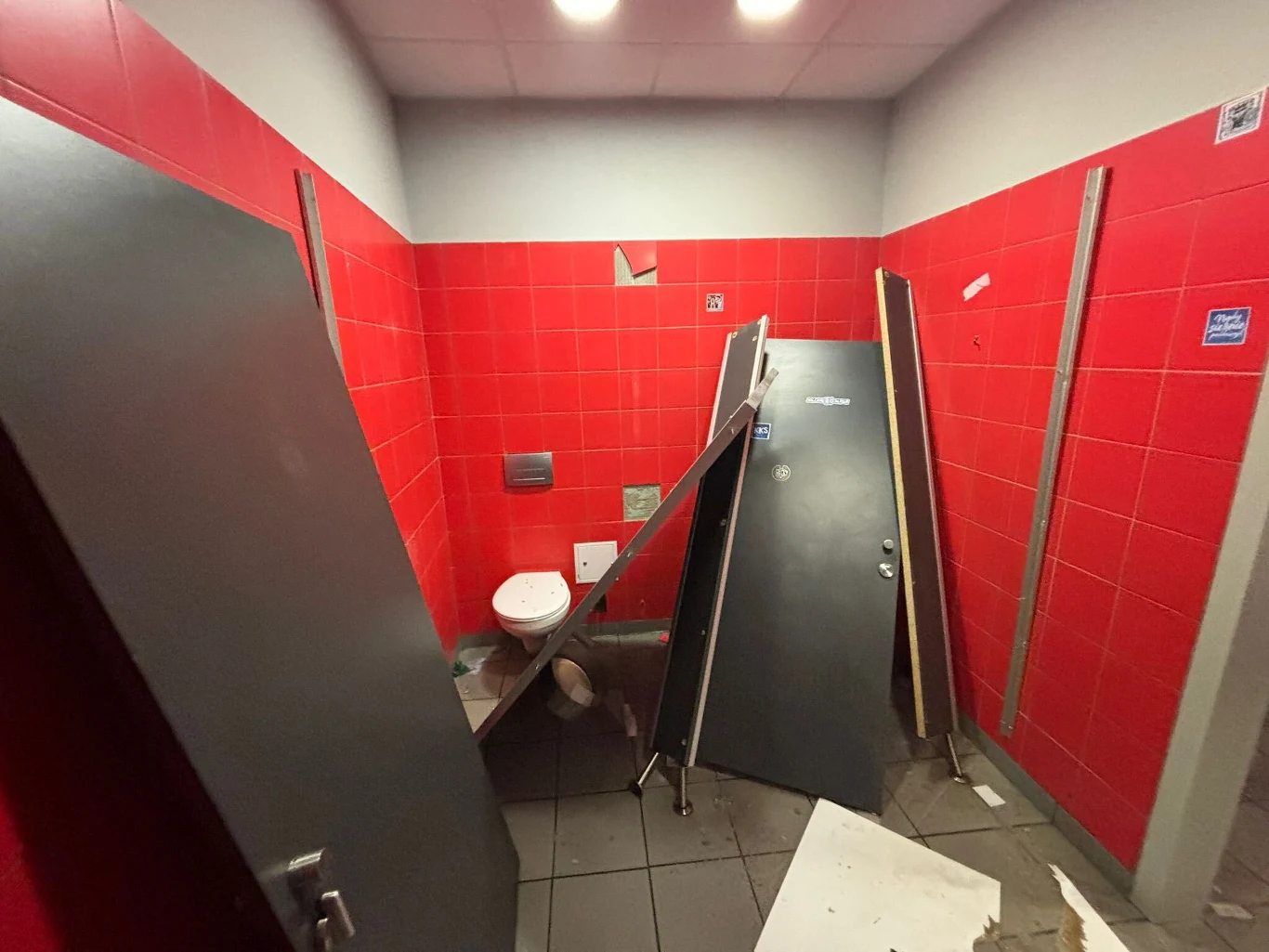 Zniszczone wnętrze publicznej toalety z czerwonymi kafelkami, przewrócona kabina, połamane drzwi i rozrzucone elementy wyposażenia, widoczna sedesowa muszla oraz ogólny bałagan i uszkodzenia.