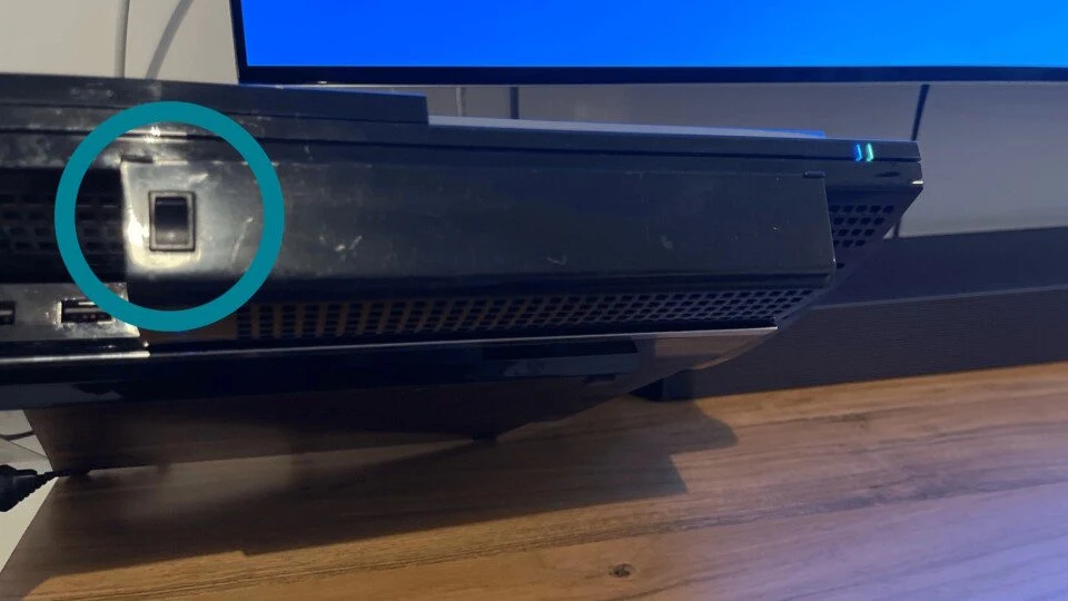 Czarny sprzęt elektroniczny z wyraźnie zaznaczonym portem optycznym, ustawiony na drewnianej powierzchni, w tle część ekranu telewizora z niebieskim wyświetlaczem.