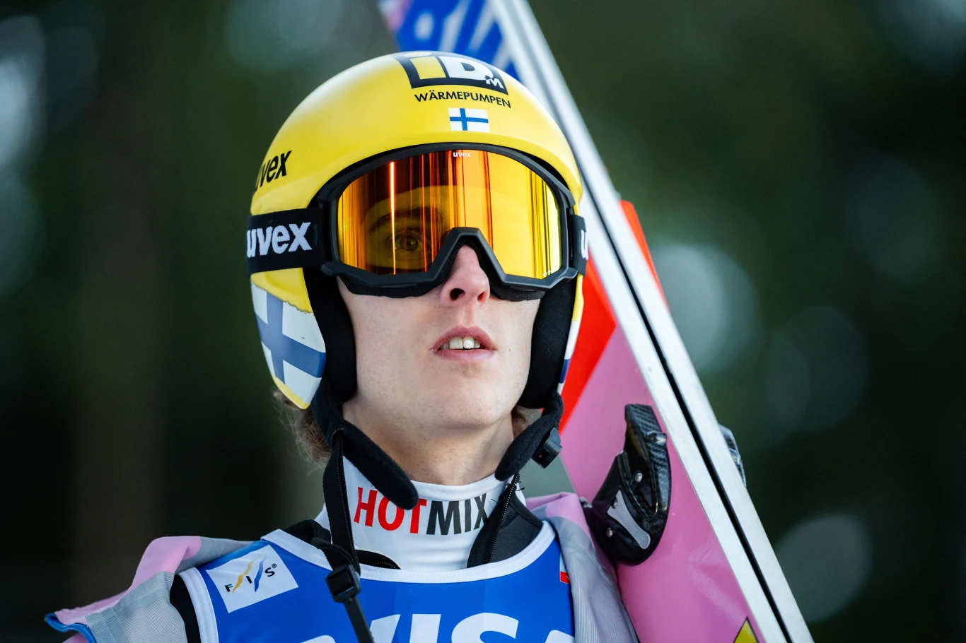 Skoczek narciarski w żółtym kasku z flagą Finlandii, ubrany w kombinezon sportowy i gogle, trzyma narty na ramieniu, skoncentrowany przed skokiem.