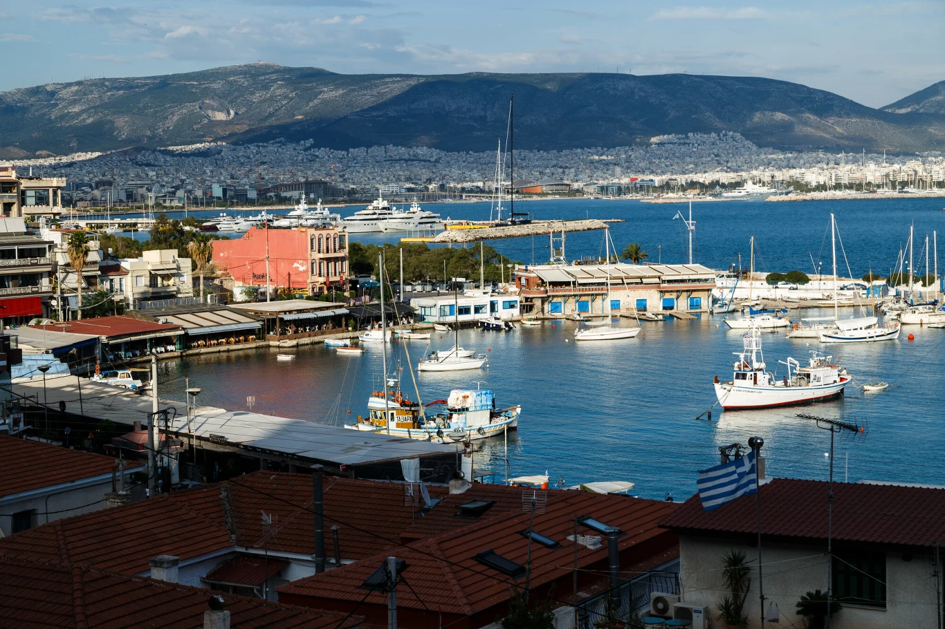Chińska mafia wykorzystywała do przemytu port w Pireus. Port w Pireus z cumującymi łodziami, jachtami, zabudową miejską i flagą Grecji w prawym dolnym rogu.