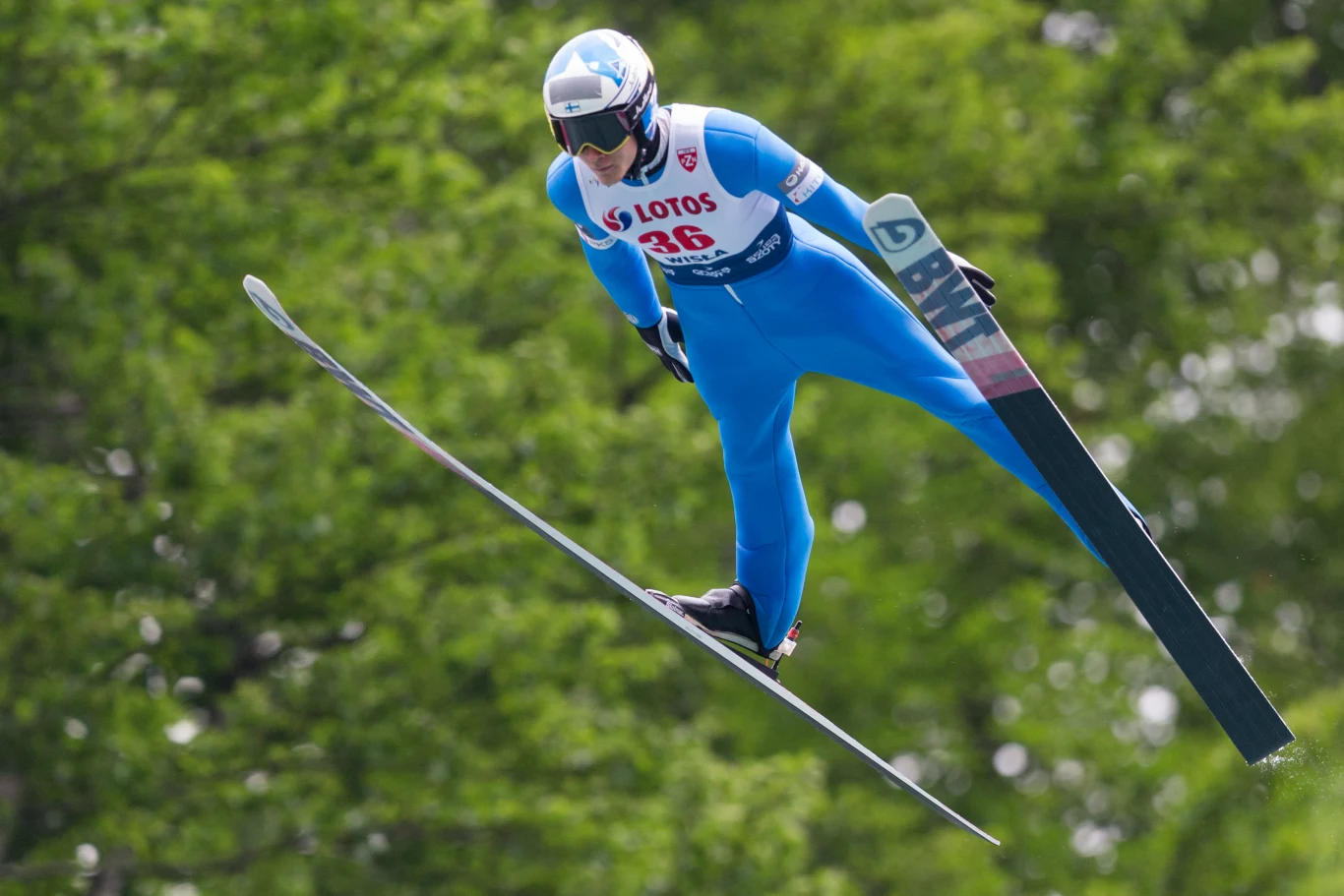 Skoczek narciarski w niebieskim kombinezonie leci nad zielonym tłem drzew, dynamicznie pochylony do przodu z szeroko rozstawionymi nartami, na kasku i stroju widoczne logotypy sponsorów.