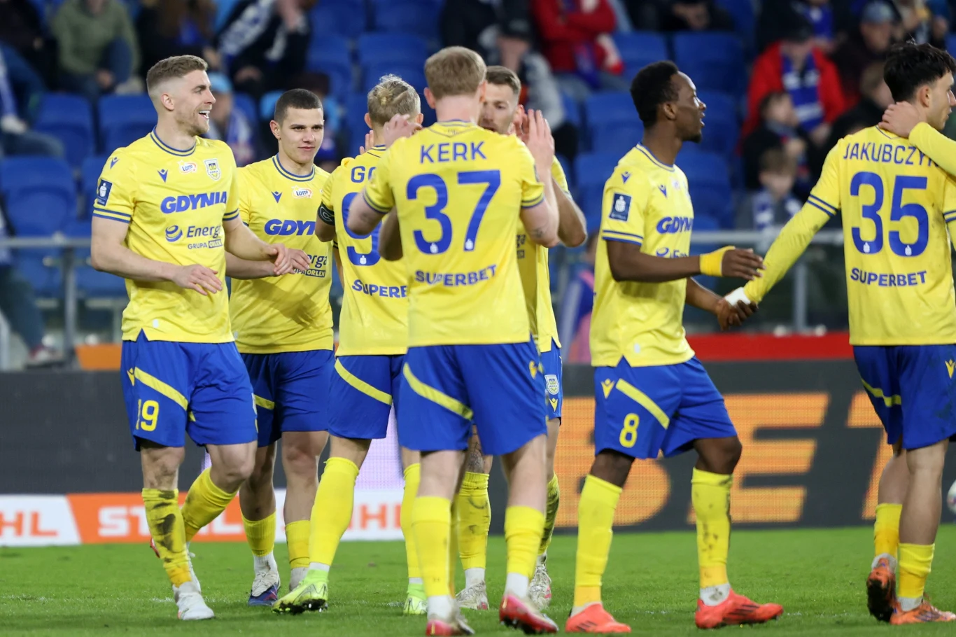 Grupa piłkarzy w żółto-niebieskich strojach drużyny Arka Gdynia świętuje wspólnie na boisku, kilku zawodników przybija sobie piątki, na dalszym planie widoczni są kibice na trybunach.