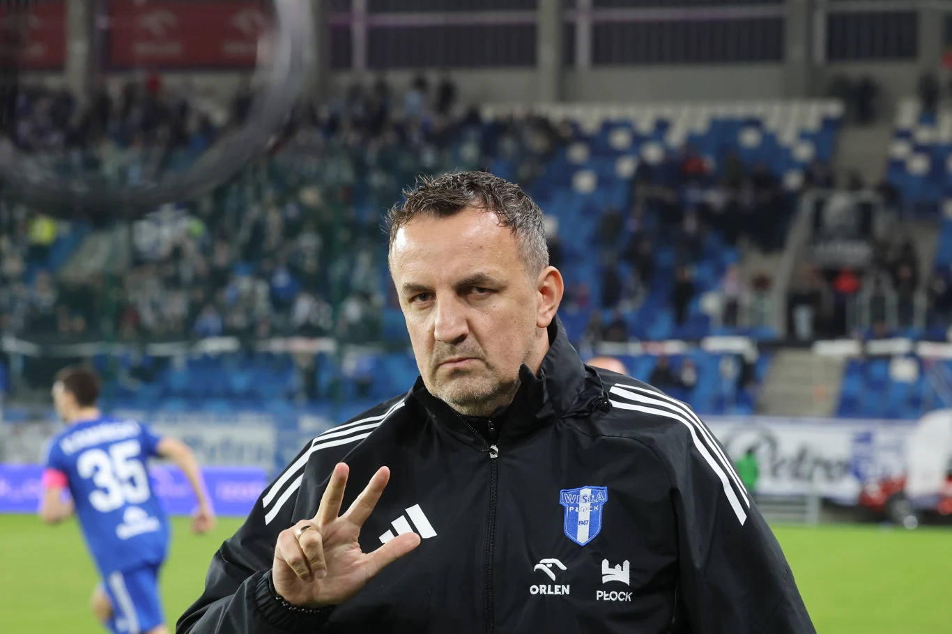Mężczyzna w czarnej sportowej kurtce z herbem klubu piłkarskiego Wisła Płock wykonuje gest trzema palcami, stojąc na tle stadionu piłkarskiego z trybunami i piłkarzami ubranymi w niebieskie stroje.