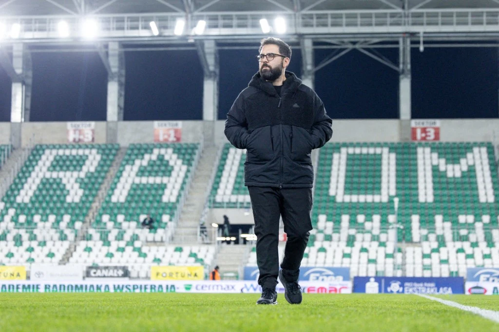 Mężczyzna w okularach i ciemnej kurtce przechadza się po murawie stadionu piłkarskiego z pustymi trybunami, na których widoczny jest napis 'RADOM'.