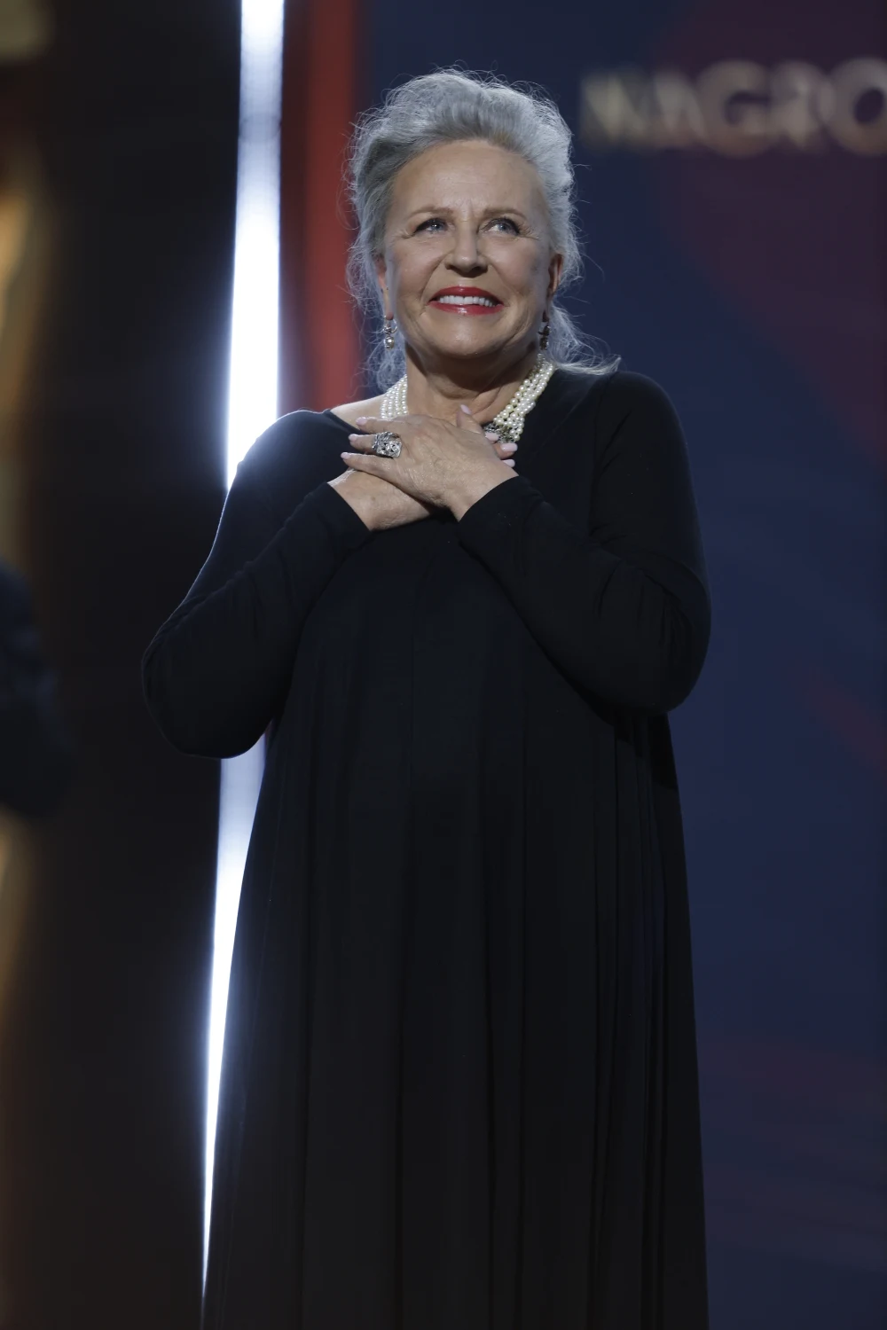 Starsza kobieta z siwymi włosami upiętymi do góry, ubrana w elegancką, czarną suknię, stoi na scenie z rękami skrzyżowanymi na piersi, z uśmiechem spoglądając przed siebie.