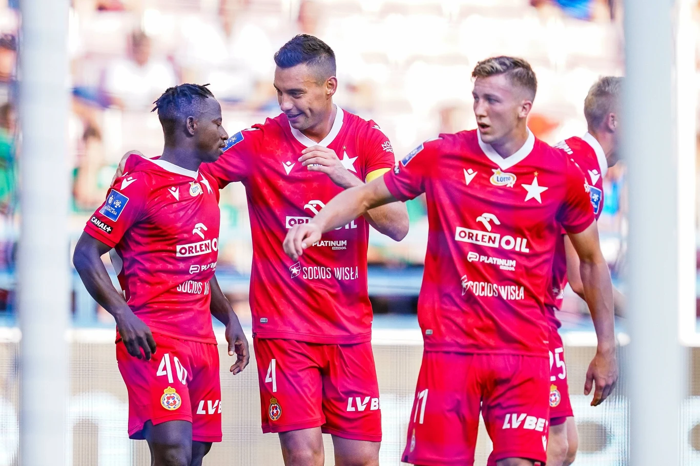 Trzech piłkarzy w czerwonych strojach drużyny Wisła Kraków świętuje na boisku, jeden z zawodników obejmuje ramieniem kolegę, wszyscy wyglądają na zadowolonych po udanej akcji podczas meczu piłkarskiego.