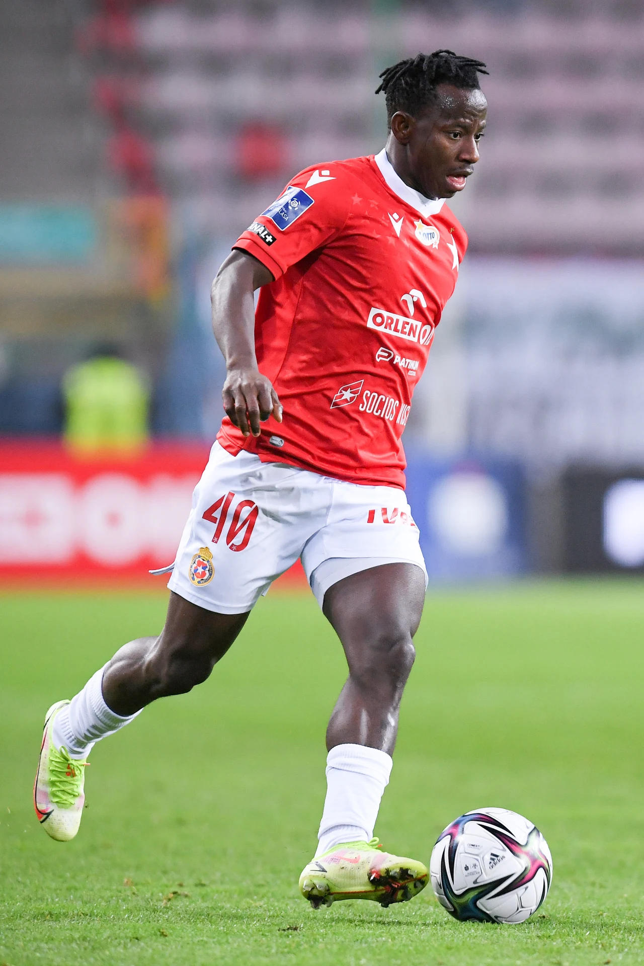 Piłkarz w czerwonej koszulce i białych spodenkach prowadzi piłkę nożną po murawie podczas meczu, koncentrując się na grze.