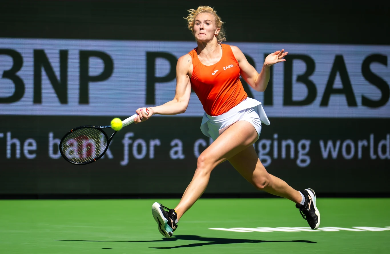 Tenisistka w pomarańczowej koszulce i białej spódniczce dynamicznie odbija piłkę podczas meczu na korcie twardym, w tle widoczne logo sponsora na bandzie reklamowej.