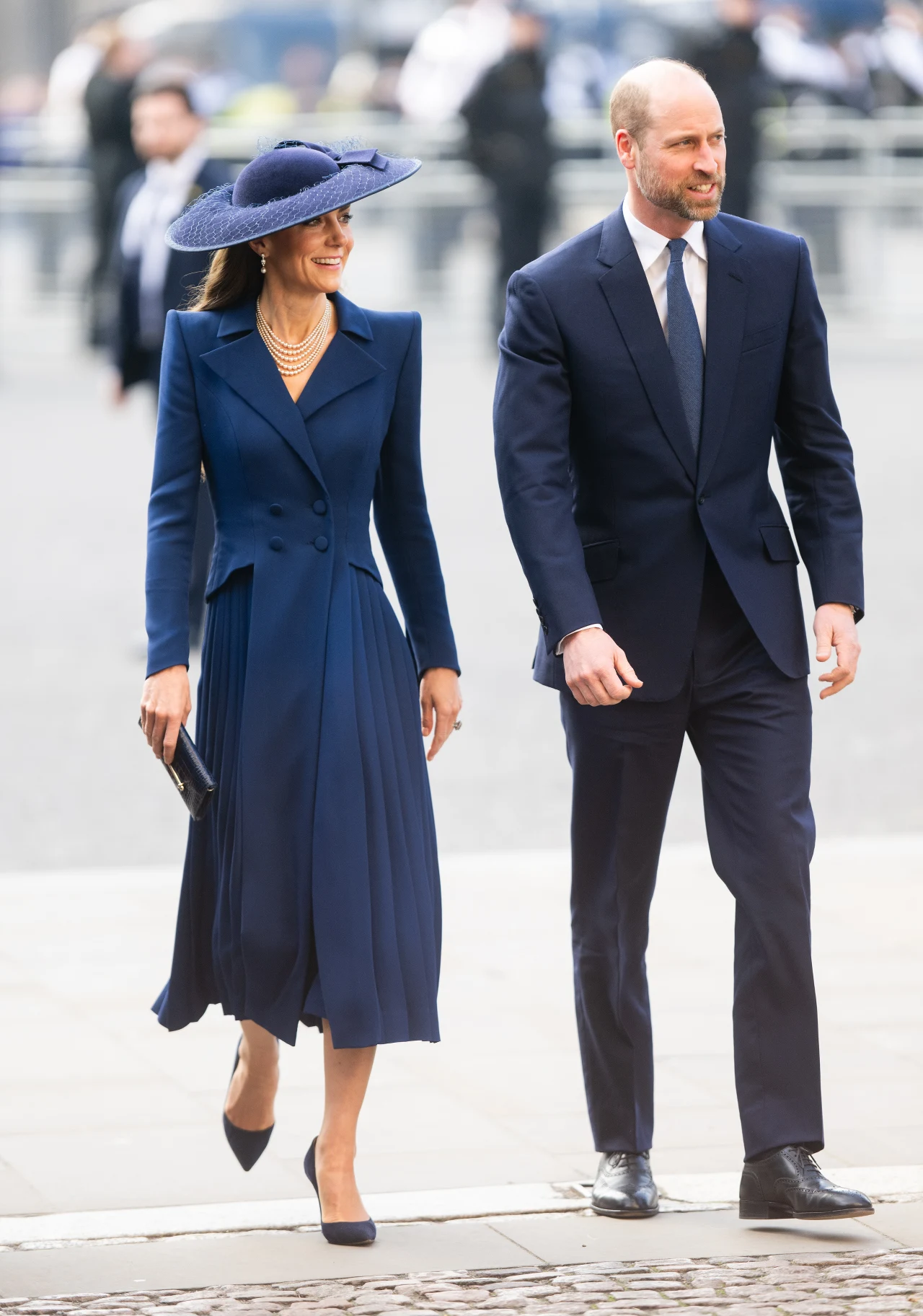 Elegancka para ubrana w granatowe stroje formalne idzie razem chodnikiem; kobieta ma na sobie długą sukienkę, szeroki kapelusz oraz perłowy naszyjnik, mężczyzna ubrany jest w ciemny garnitur i krawat.