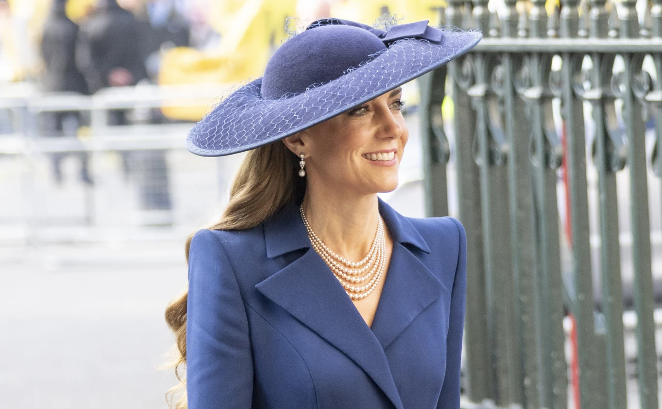 Elegancko ubrana kobieta w niebieskim kapeluszu z szerokim rondem, w dopasowanym płaszczu w tym samym kolorze, ozdobiona perłowym naszyjnikiem i kolczykami, uśmiecha się i idzie obok metalowego ogrodzenia.