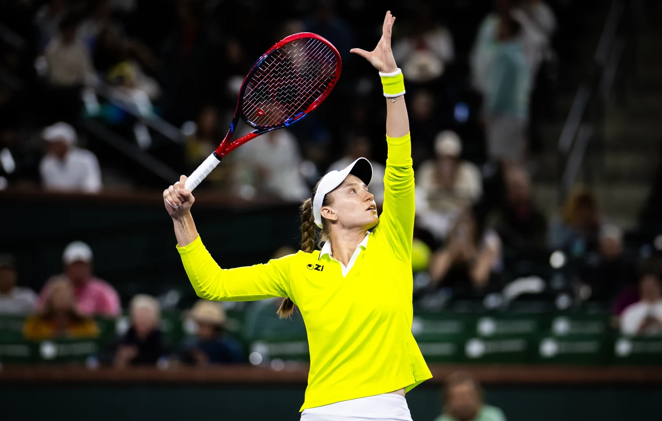 Tenisistka w żółtej bluzie i białej czapce wykonuje serwis podczas meczu, trzymając rakietę w jednej ręce i unosząc drugą do góry; na trybunach widać rozmytych kibiców.