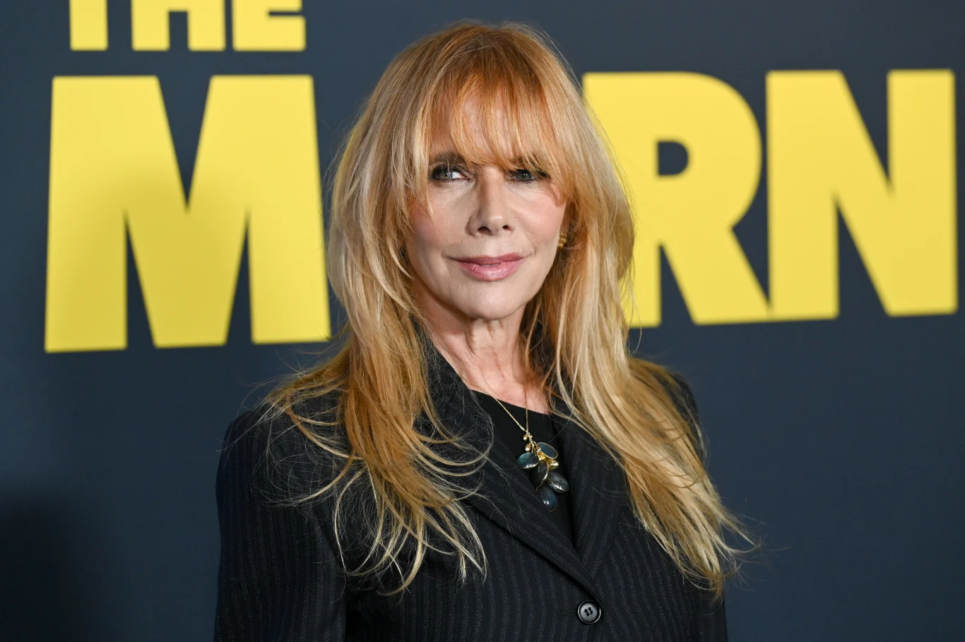 Kobieta o długich blond włosach w eleganckim czarnym garniturze, stojąca na tle czarno-żółtej ścianki z dużym napisem.