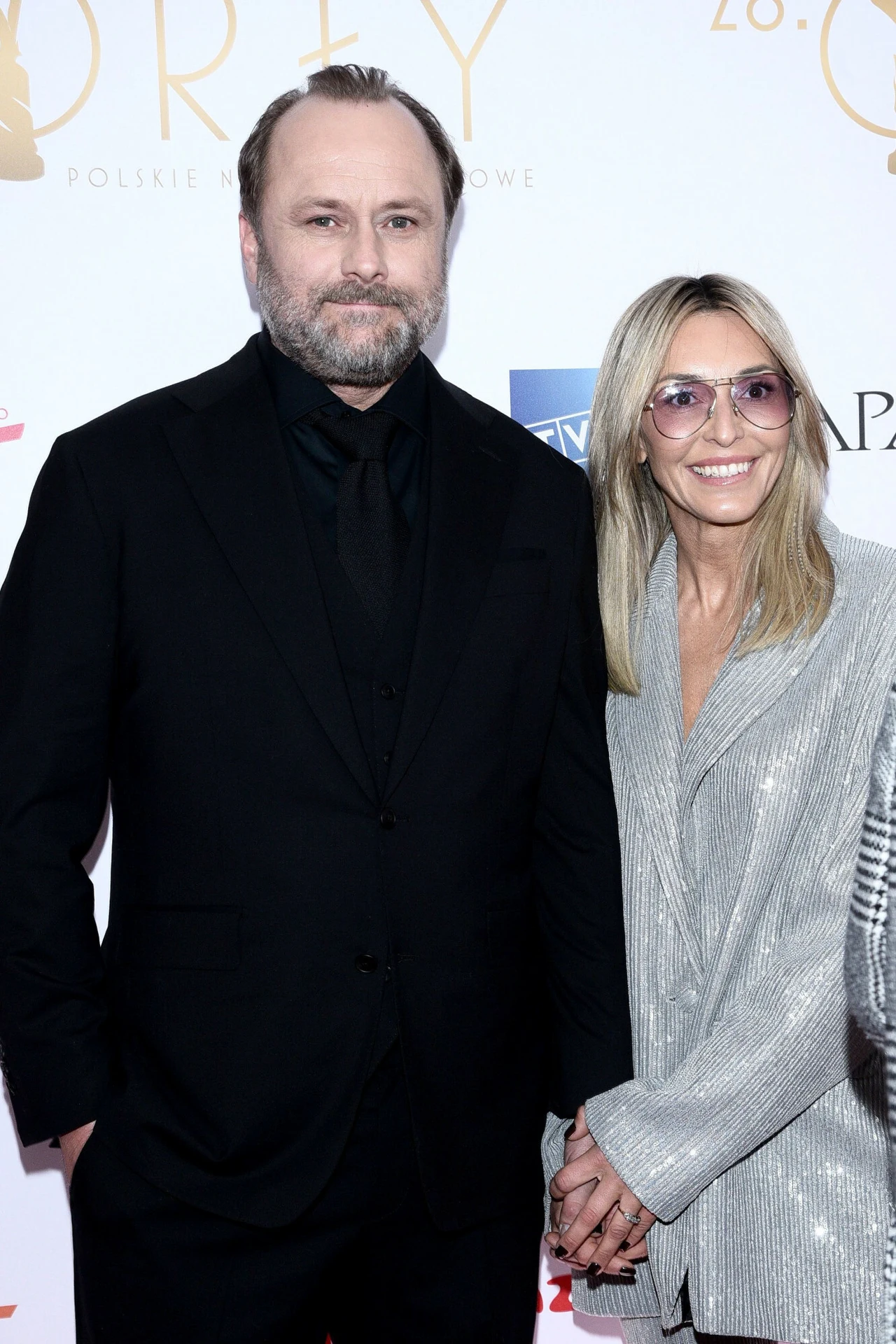 Mężczyzna w czarnym garniturze stoi obok uśmiechniętej kobiety w srebrzystej marynarce i okularach, na tle ścianki z logotypami podczas oficjalnego wydarzenia.