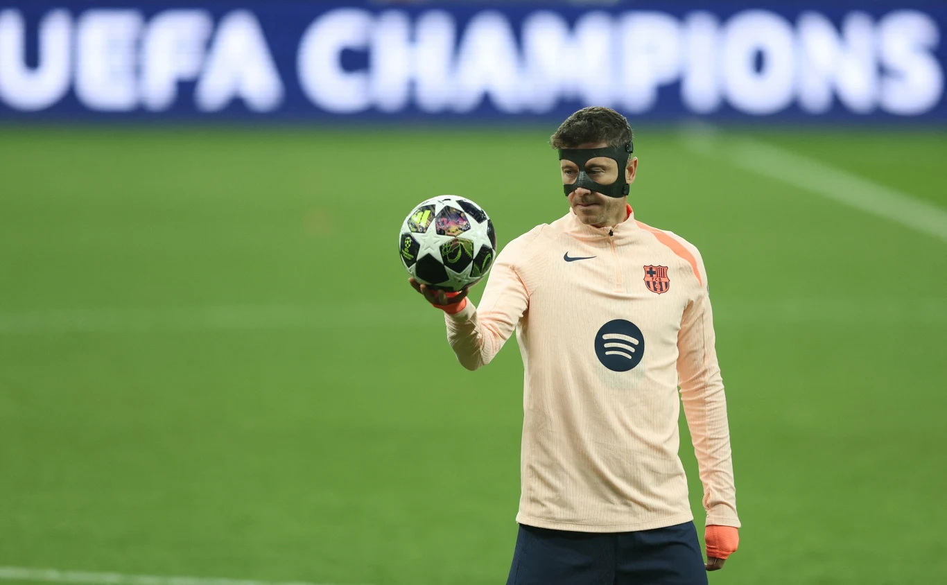 Piłkarz Barcelony w jasnej bluzie treningowej i czarnej masce na twarzy trzyma piłkę Ligi Mistrzów UEFA, stojąc na boisku na tle rozmytego napisu 'UEFA CHAMPIONS'.