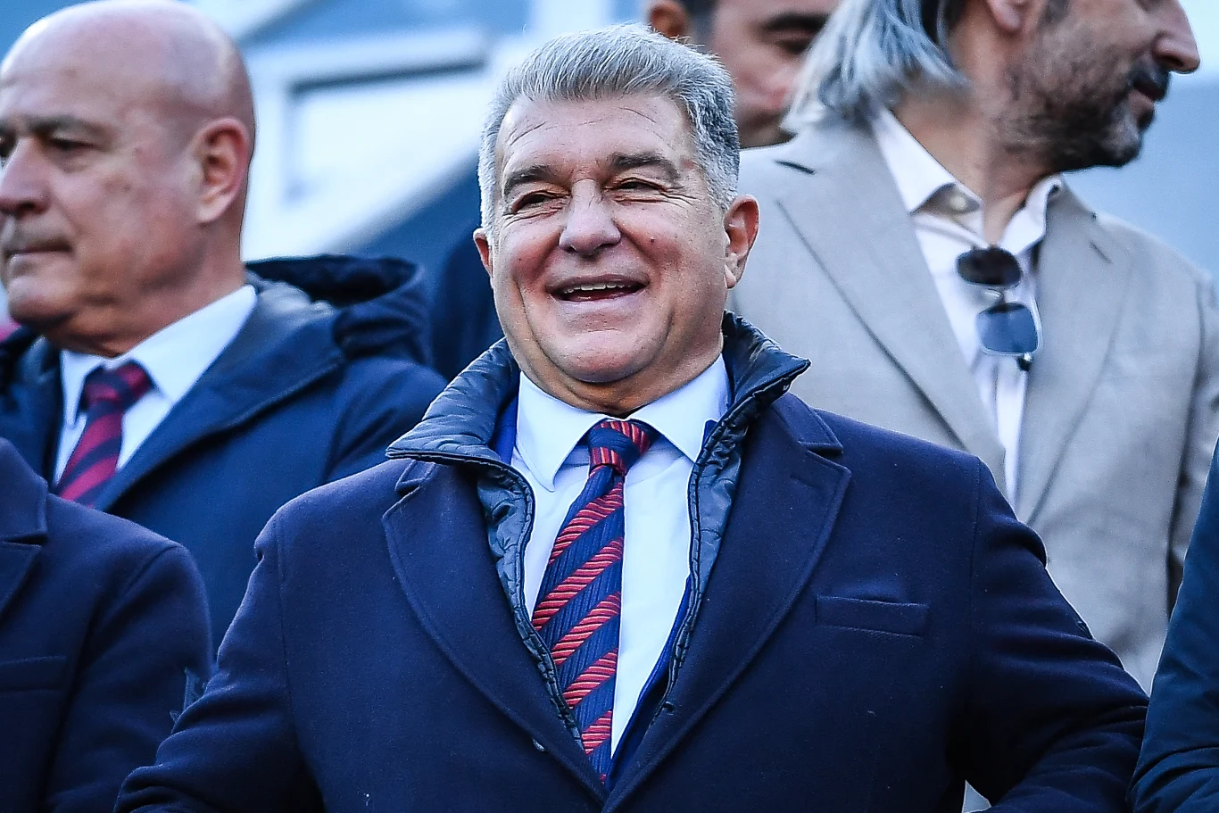 Uśmiechnięty mężczyzna w garniturze i krawacie w granatowo-czerwone paski stoi wśród innych elegancko ubranych osób, dominują ciemne kolory ubrań.