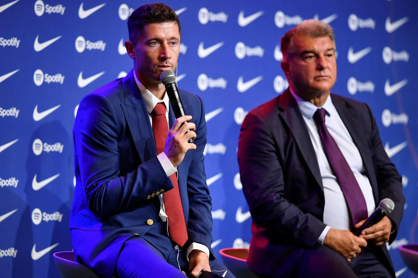 Dwóch mężczyzn w garniturach siedzi na scenie w trakcie konferencji prasowej, jeden z nich trzyma mikrofon i mówi, w tle ścianka z logotypami Spotify i Nike.