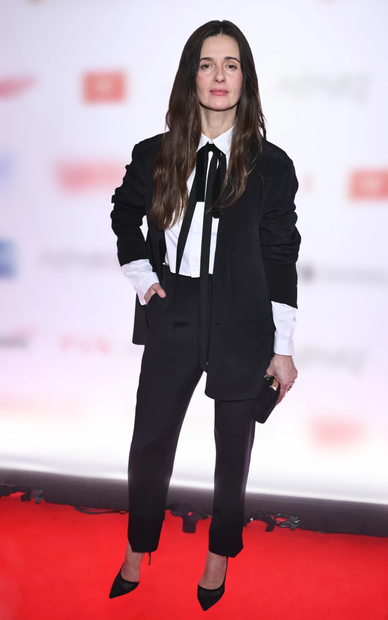 Kobieta z długimi, ciemnymi włosami ubrana w elegancki, czarny garnitur z białą koszulą i czarną wąską kokardą pod szyją, stoi na czerwonym dywanie, trzymając w dłoni małą czarną kopertówkę; tło rozmyte, z widocznymi czerwonymi i szarymi napisami.