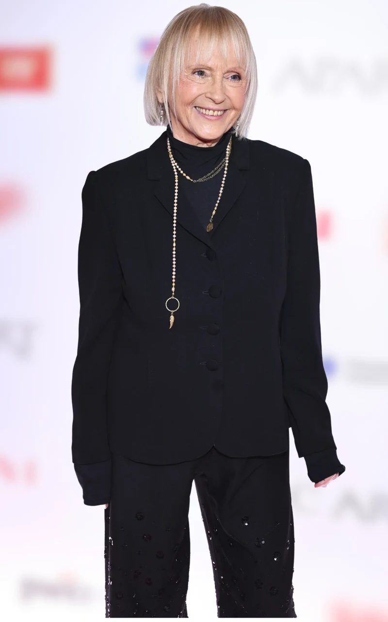 Uśmiechnięta kobieta z blond włosami do ramion ubrana w elegancki czarny garnitur i czarny golf, ozdobiona długim, złotym naszyjnikiem, stoi na tle jasnej ścianki z niewyraźnymi logotypami w tle.