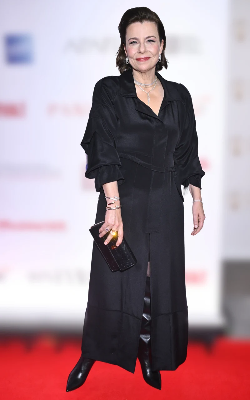 Kobieta w eleganckiej, czarnej sukni do kostek i czarnych, lakierowanych butach, trzymająca kopertówkę i ozdobiona delikatną biżuterią, stoi na czerwonym dywanie przed ścianą z logotypami.