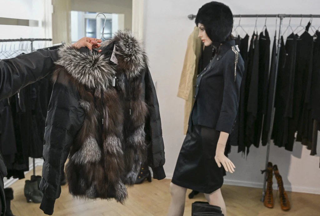 Czarna kurtka z dużym, puszystym kołnierzem z futra trzymana na wieszaku, w tle manekin ubrany w czarną spódnicę, żakiet i futrzaną czapkę w sklepie odzieżowym z eleganckimi ubraniami na wieszakach.