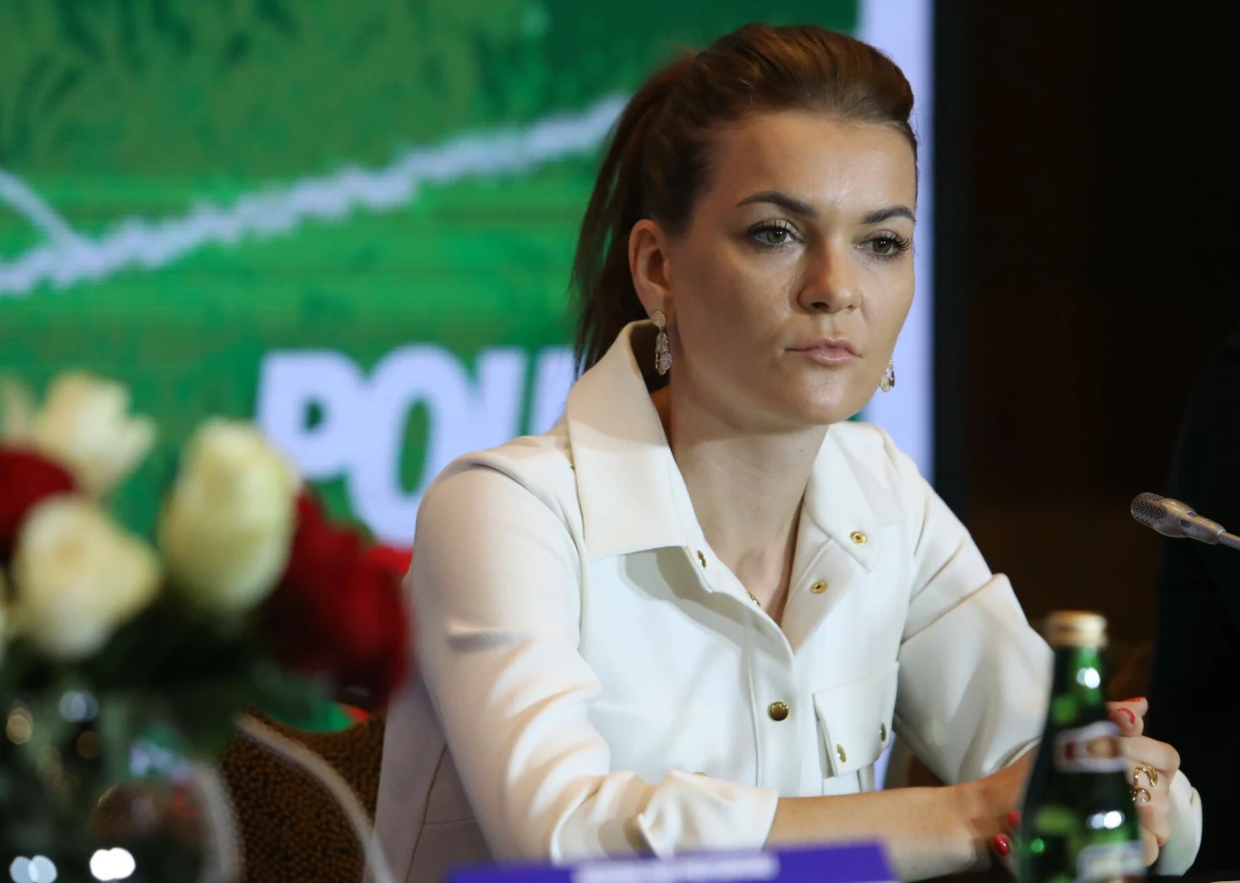 Kobieta z upiętymi włosami w białej koszuli siedzi przy stole konferencyjnym, na którym znajduje się butelka wody oraz mikrofon, w tle zielony baner z częściowo widocznym napisem.