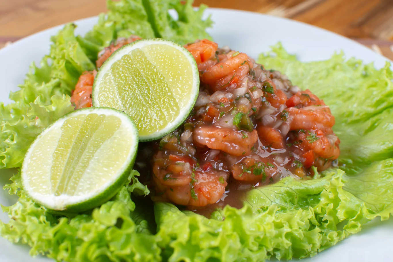 Krewetki w sosie z ziołami i drobno pokrojonymi warzywami podane na liściach świeżej sałaty, ozdobione plasterkami limonki.