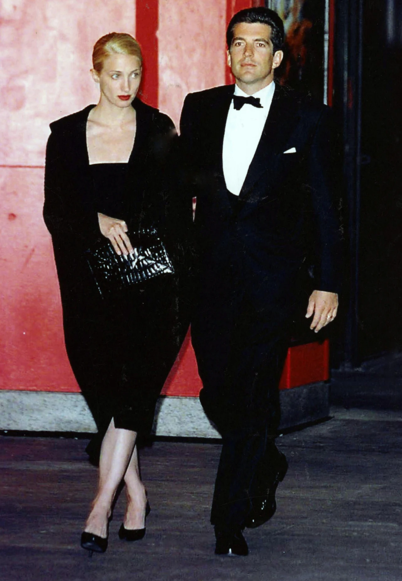 Elegancko ubrana para idzie razem, kobieta w czarnej sukni i z torebką, mężczyzna w czarnym smokingu, ciemne tło z czerwonymi akcentami.