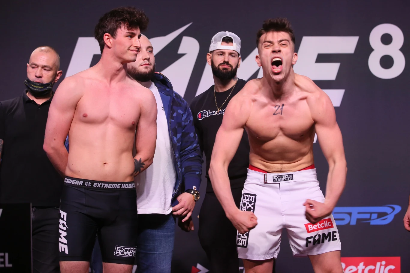 Dwóch mężczyzn w szortach sportowych stoi na scenie podczas oficjalnej ceremonii ważenia przed walką, jeden z nich prezentuje umięśnione ciało i wykonuje bojową pozę z zaciśniętymi pięściami oraz krzykiem, podczas gdy drugi zachowuje spokojną postawę i...