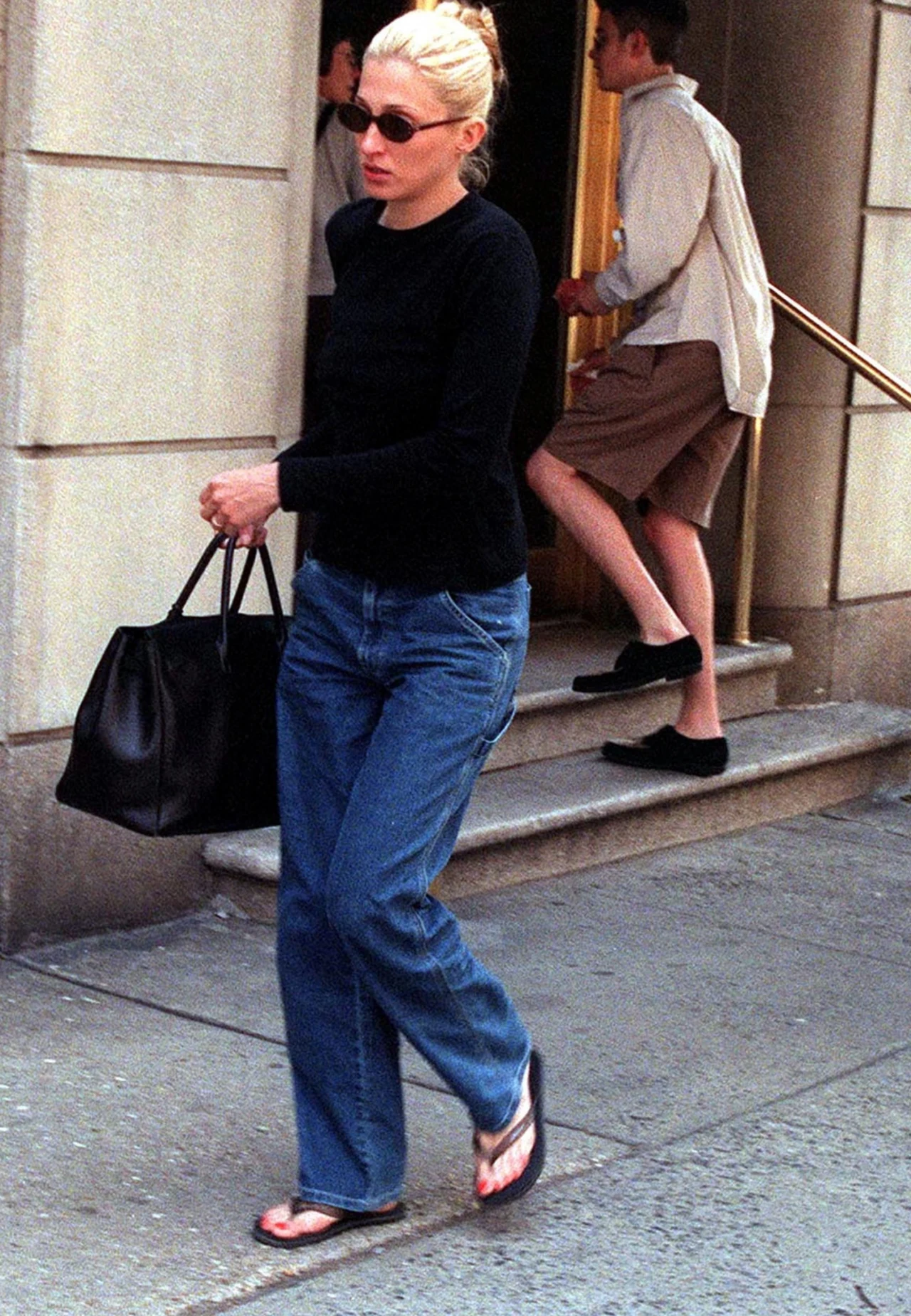 Kobieta w czarnych okularach przeciwsłonecznych, czarnym swetrze i niebieskich dżinsach spaceruje chodnikiem, niosąc dużą czarną torbę, ubrana w japonki i mająca związaną blond fryzurę.