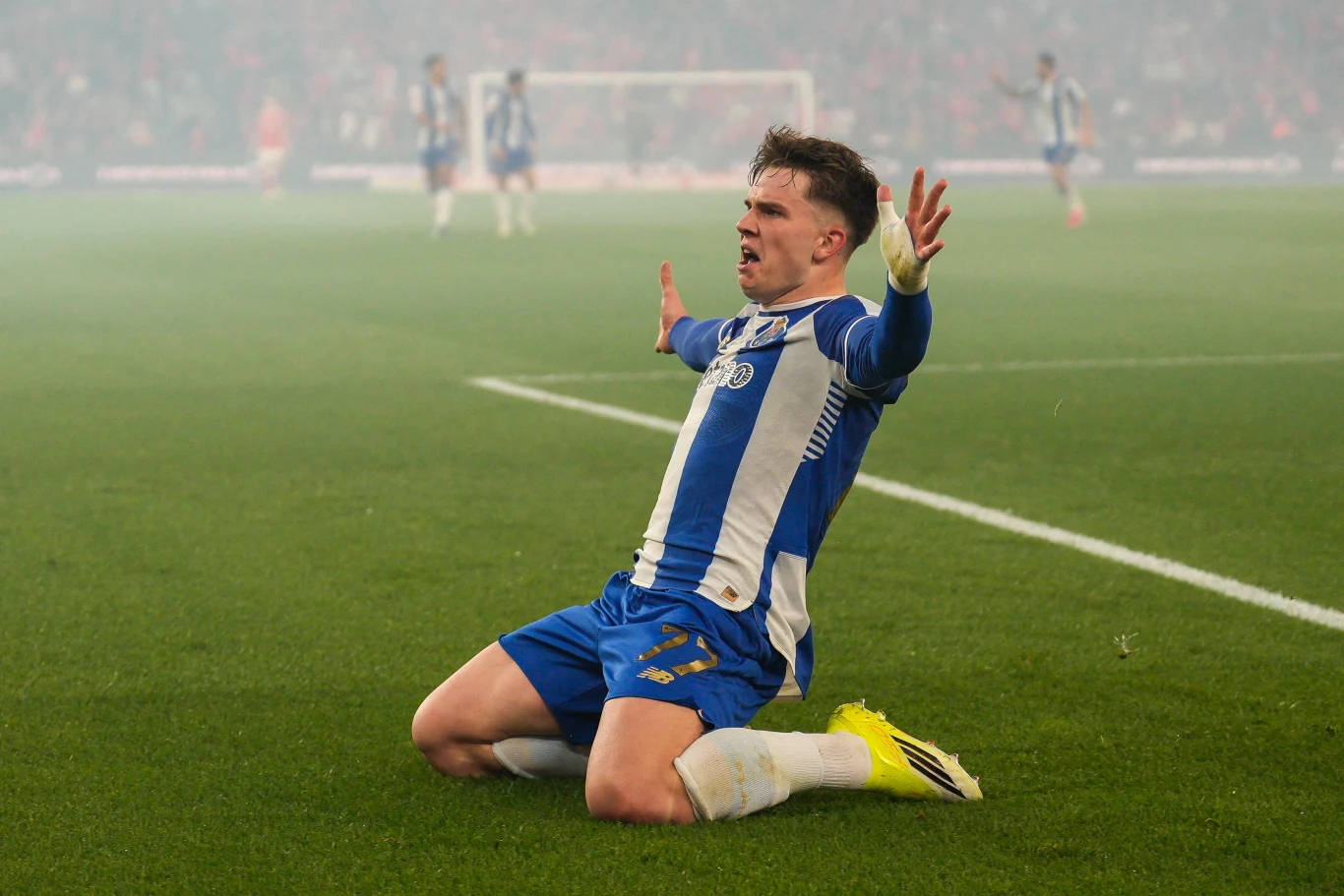 Piłkarz w niebiesko-białym stroju świętuje zdobycie gola, klęcząc na murawie z rozłożonymi ramionami, wokół rozległe boisko i niewyraźna sylwetka bramki w tle.