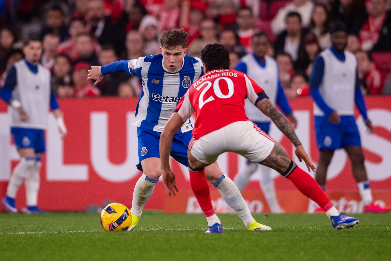 Dwóch piłkarzy rywalizuje o piłkę podczas meczu; jeden zawodnik ubrany w biało-niebieski strój FC Porto walczy z przeciwnikiem w czerwonym stroju z numerem 20, wokół nich widoczna jest rozmazana publiczność i ławka rezerwowych.