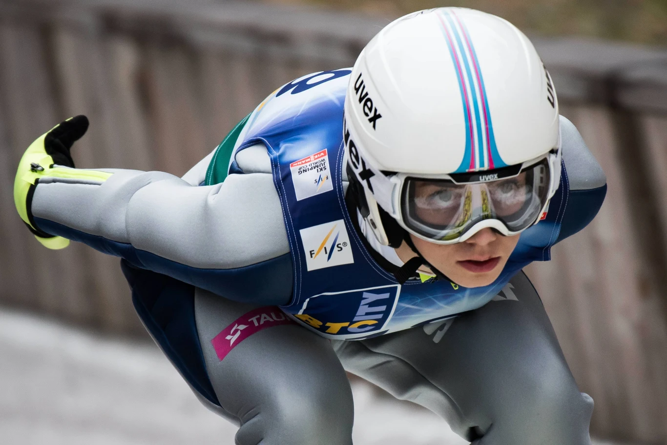 Skoczek narciarski w aerodynamicznej pozycji do lądowania, ubrany w kombinezon sportowy, kask i gogle, skoncentrowany podczas wykonywania skoku na skoczni narciarskiej.