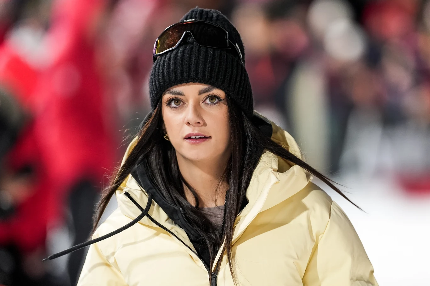 Kobieta w żółtej kurtce zimowej i czarnej czapce z okularami przeciwsłonecznymi na czubku głowy stoi na tle rozmytego tłumu ubranych na czerwono ludzi.