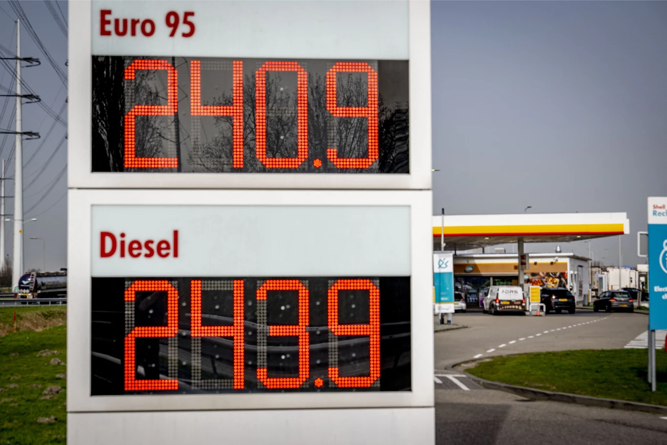 Elektroniczny wyświetlacz cen paliw na stacji benzynowej pokazuje aktualne ceny Euro 95 i diesla, obie wynoszą 240,9; w tle widoczne stanowiska do tankowania oraz zaparkowane samochody.