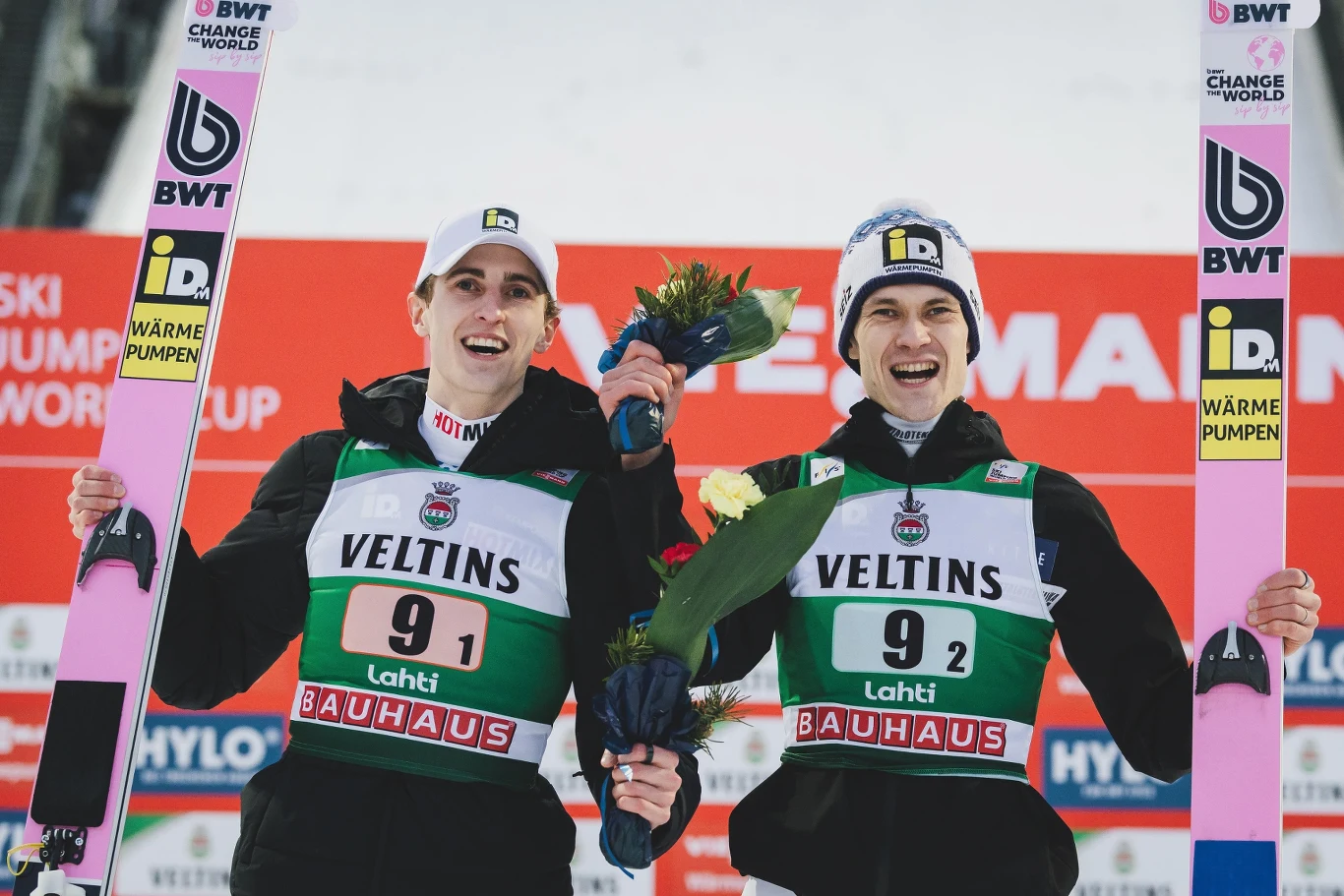 Dwóch uśmiechniętych skoczków narciarskich w sportowych strojach i numerami startowymi, trzymających narty oraz bukiety kwiatów, stojących na tle tablicy z logo sponsorów po zakończonych zawodach z cyklu Pucharu Świata.