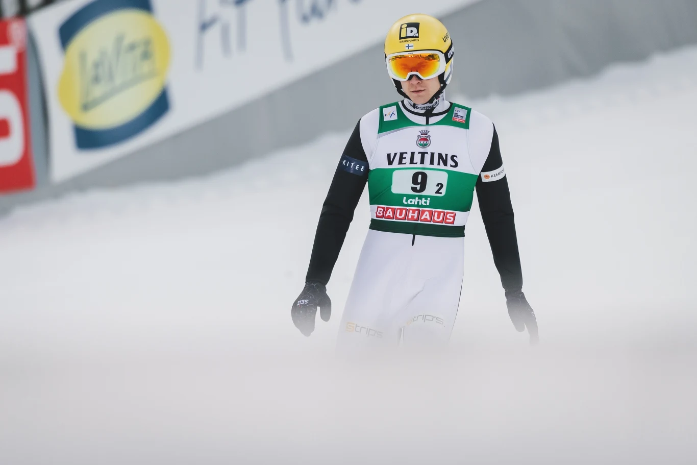 Skoczek narciarski w kombinezonie z numerem 92 i żółtym kaskiem stoi na zaśnieżonym zeskoku, otoczony lekką mgłą, z widocznymi reklamami w tle.