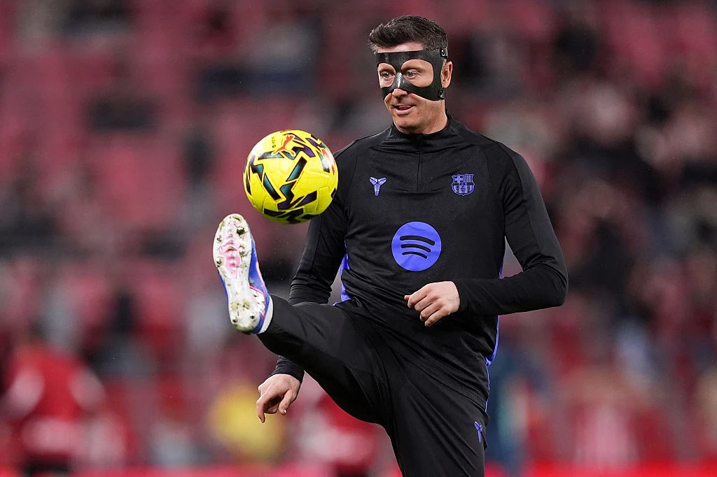 Robert Lewandowski Robert Lewandowski w czarnej masce ochronnej żongluje żółto-czarną piłką podczas rozgrzewki na stadionie.