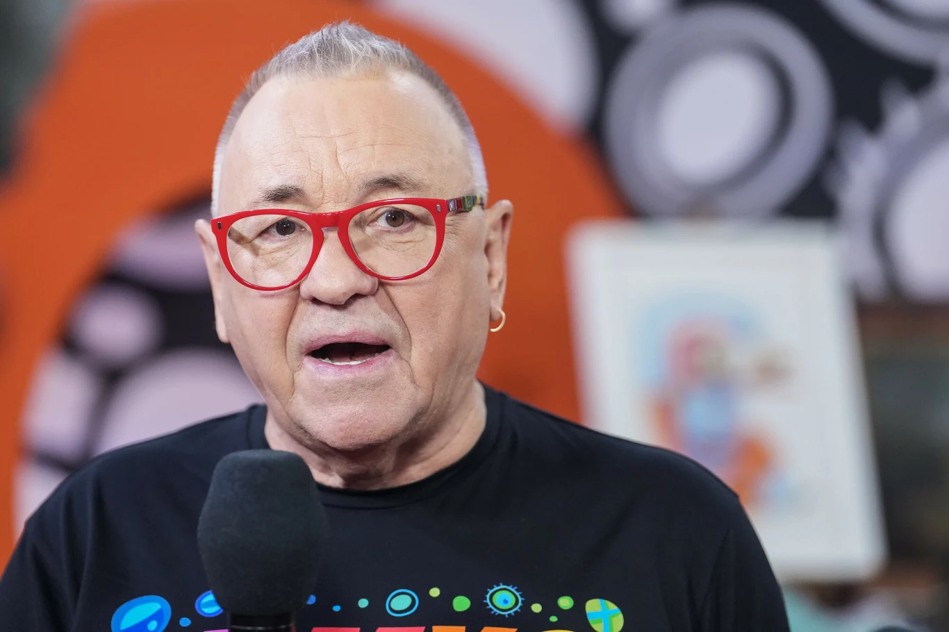 Jurek Owsiak ogłosił kolejnego wykonawcę Pol'and'Rock Festivalu Jurek Owsiak w okularach z czerwoną oprawką, z mikrofonem, na tle wyrazistej grafiki podczas ogłoszenia wykonawcy Pol'and'Rock Festivalu.