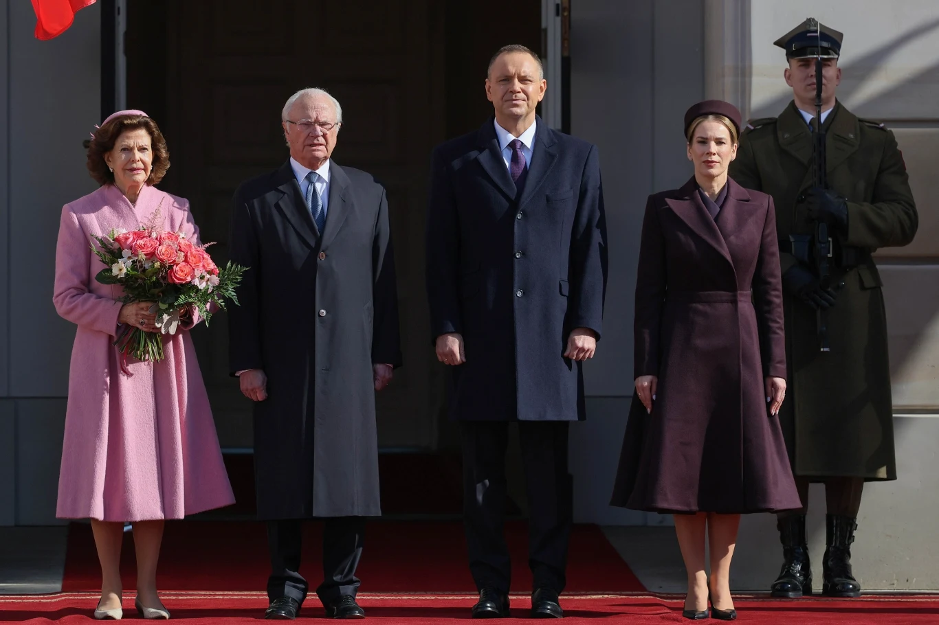 Cztery elegancko ubrane osoby stoją na czerwonym dywanie przed wejściem do budynku; kobieta po lewej trzyma bukiet kwiatów, obok stoi mężczyzna w okularach, obok niego kolejny mężczyzna oraz kobieta ubrana na ciemno, po prawej stronie stoi żołnierz w m...