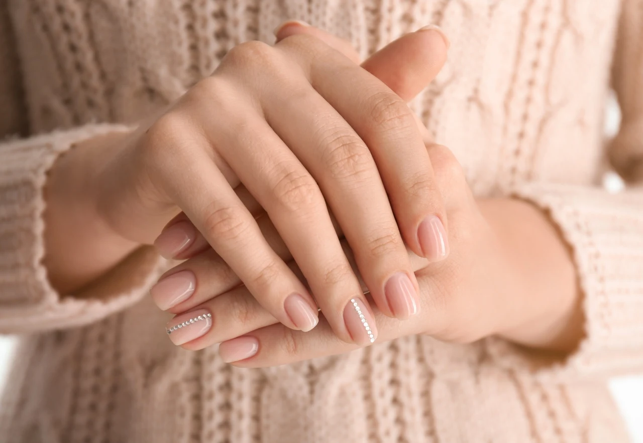 Dłonie z zadbanym manicure, paznokcie pomalowane jasnoróżowym lakierem z delikatnymi ozdobami w formie drobnych białych kropek na jednym z palców, tło stanowi sweter o beżowym odcieniu z wyraźnie widoczną fakturą dzianiny.