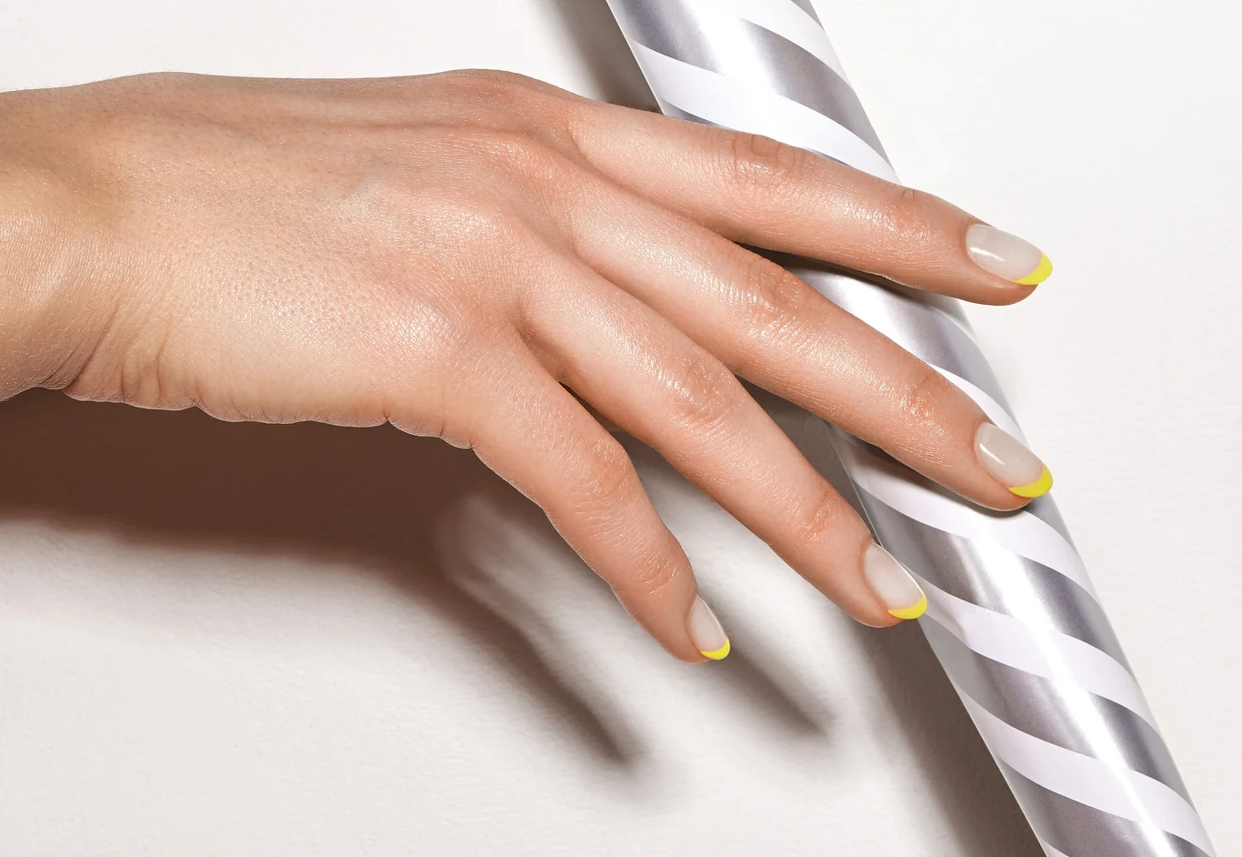 Dłoń z zadbanymi paznokciami, na których widoczna jest delikatna stylizacja french manicure z żółtymi końcówkami, oparta o biało-srebrny, spiralny element.