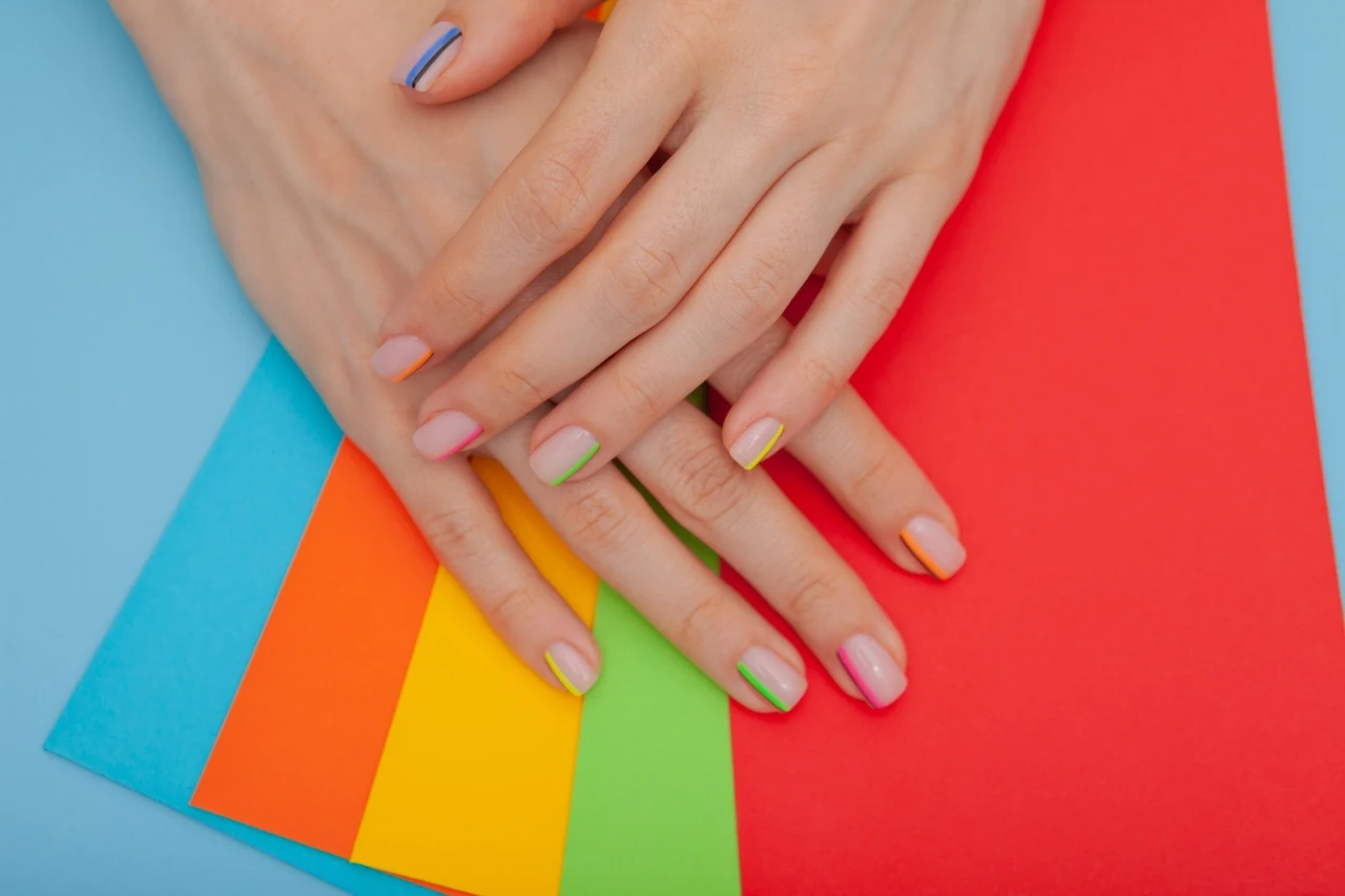 Zadbane dłonie z paznokciami pomalowanymi na pastelowe kolory, spoczywające na zestawie kolorowych kartek w odcieniach niebieskiego, pomarańczowego, żółtego, zielonego i czerwonego.