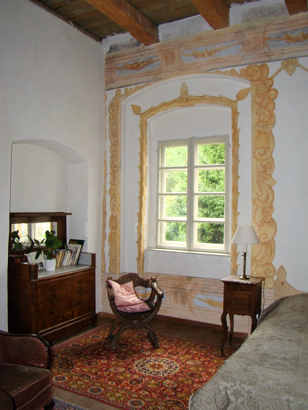 Pokój urządzony w stylu klasycznym z dużym oknem ozdobionym sztukateriami, zabytkowymi meblami z drewna, orientalnym dywanem, lampą stołową oraz roślinami w doniczkach na kredensie.