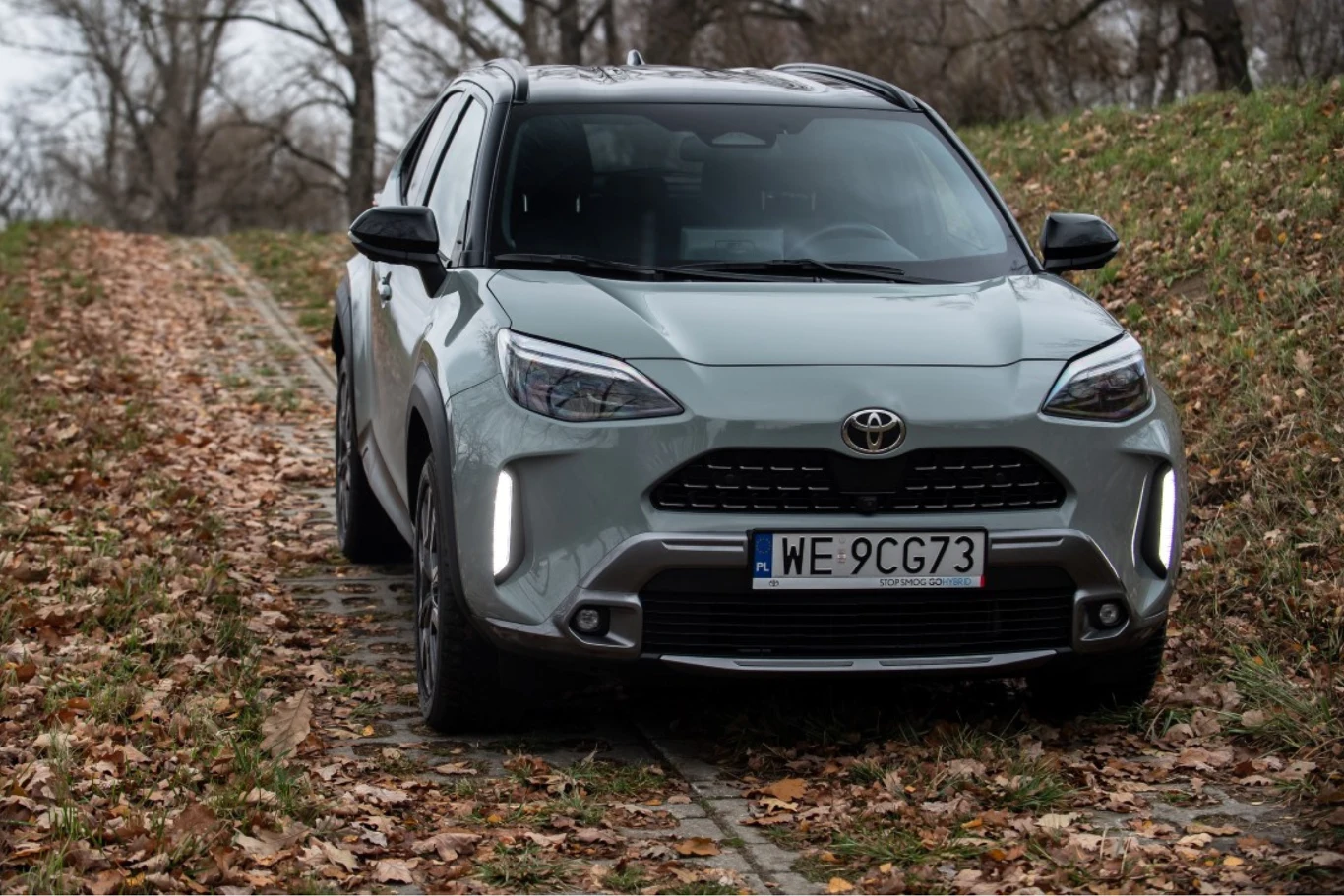 To najbardziej przystępny samochód w gamie Toyoty z napędem 4x4. Najtańsza Toyota z napędem 4x4, srebrny Yaris Cross na betonowej drodze wśród drzew bez liści.