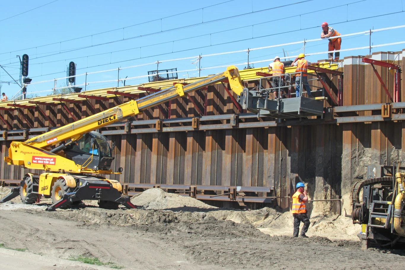 Ankieta: 4 na 10 firm planuje rekrutacje w drugim kwartale 2026 r. Pracownicy budowlani w kaskach montują elementy na rusztowaniu przy torach kolejowych z podnośnika koszowego. Pracują w słońcu.