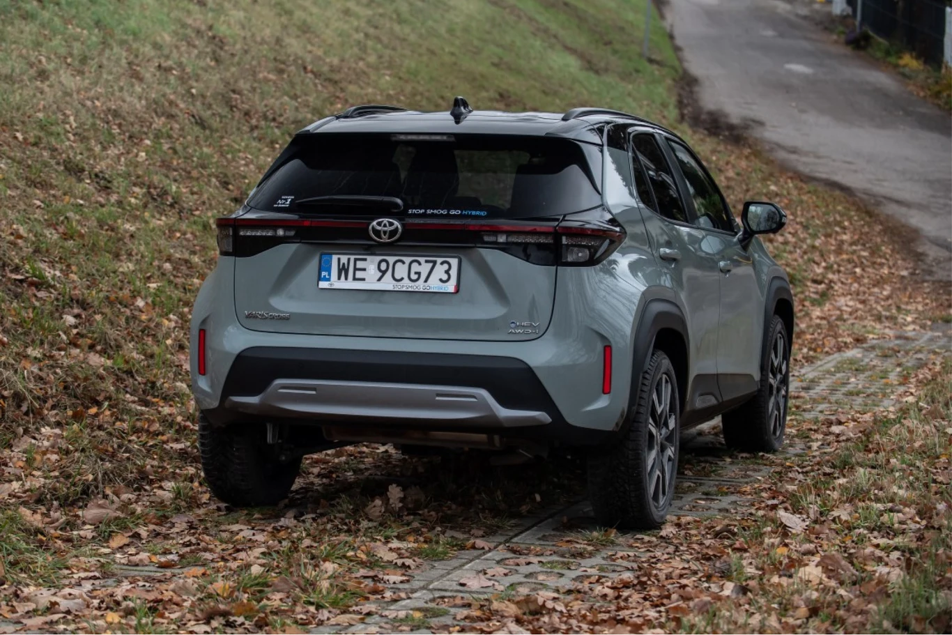 Hybrydowy układ napędowy generuje moc 130 KM. Srebrny SUV marki Toyota z widoczną rejestracją na polskich numerach, zaparkowany na utwardzonej drodze wśród opadłych, jesiennych liści i trawy.