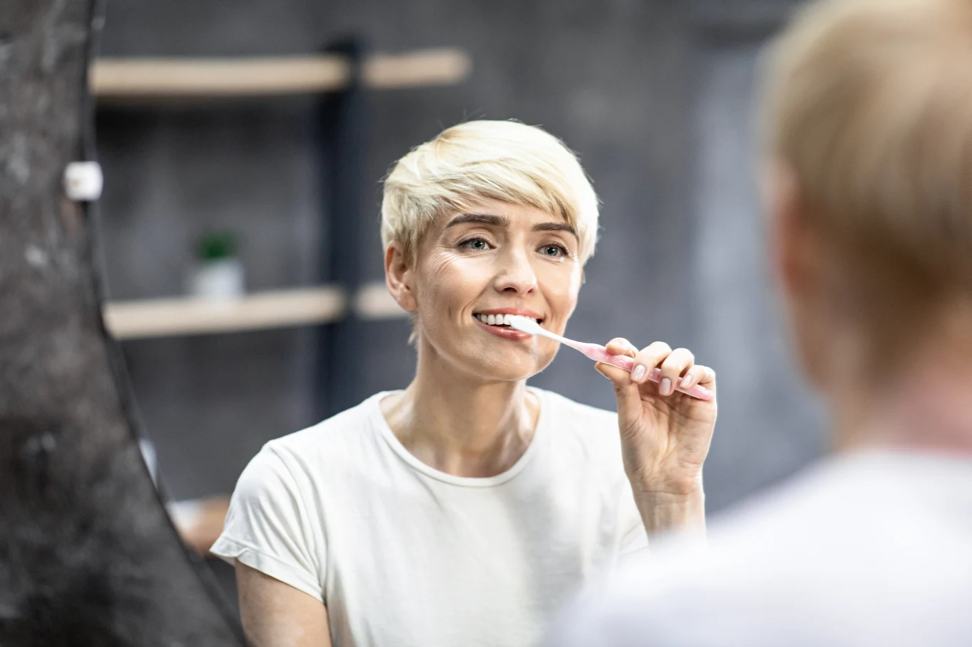 Kobieta o krótkich blond włosach czyści zęby szczoteczką przed lustrem, uśmiecha się lekko i nosi białą koszulkę.